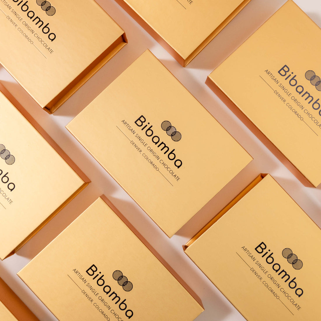 Multiple gold-colored rectangular boxes of Bibamba artisan single-origin chocolate arranged diagonally on a white background.