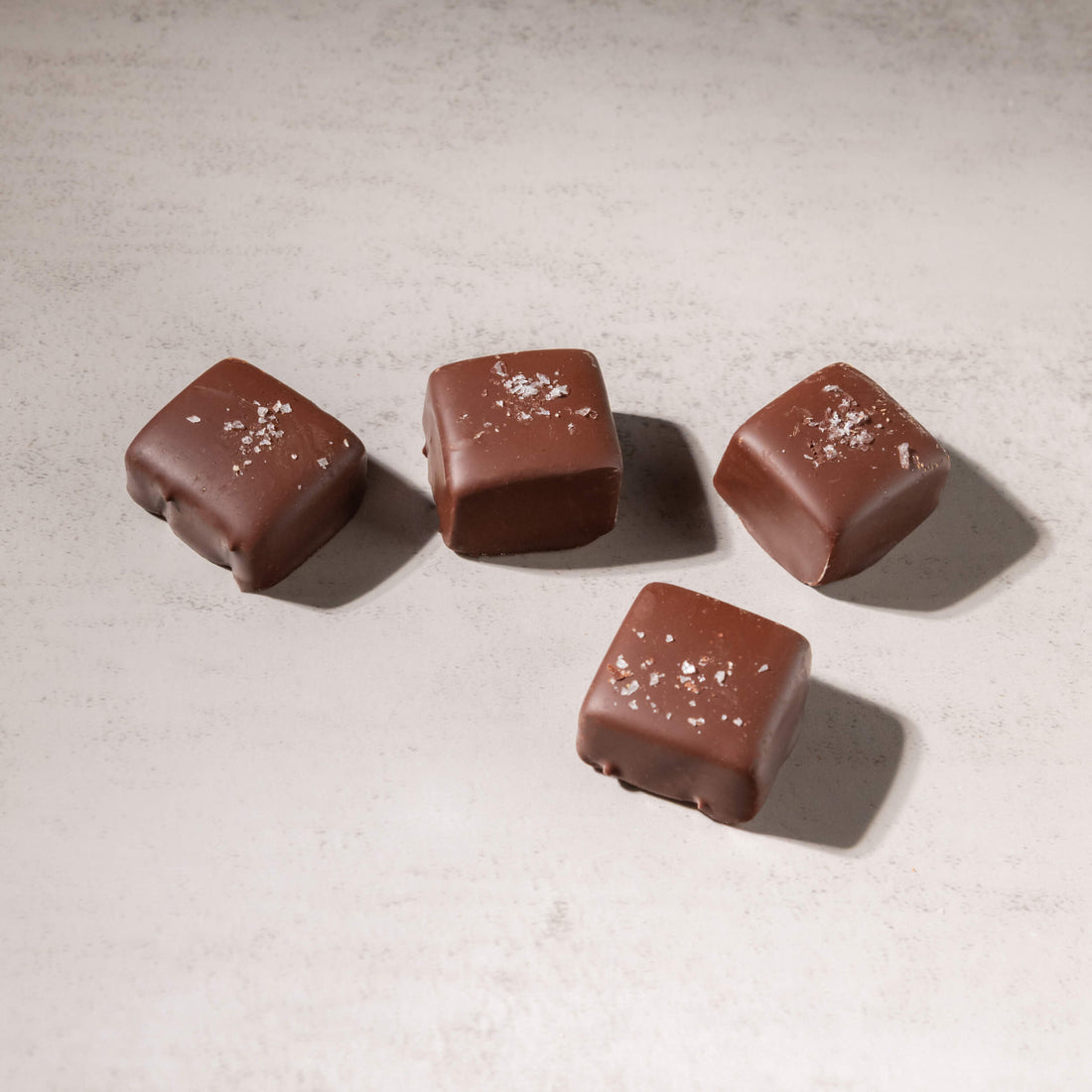 Four square chocolate candies with a glossy milk-chocolate coating and a sprinkle of coarse sea salt on top, arranged on a light textured surface.