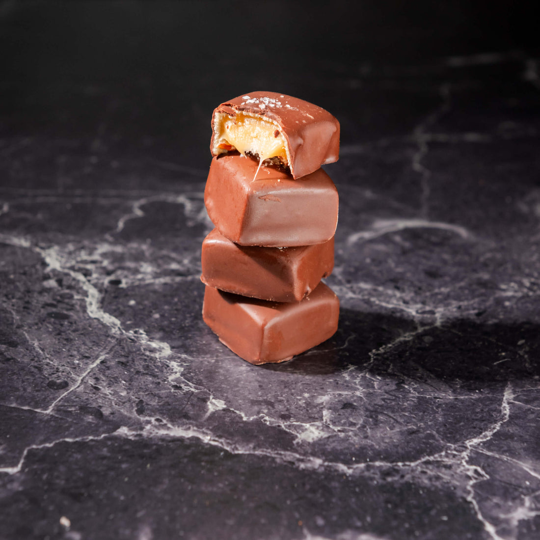 Four Bibamba milk-chocolate caramel squares stacked vertically on a dark marble surface, with the top piece broken in half to reveal a caramel-like filling.