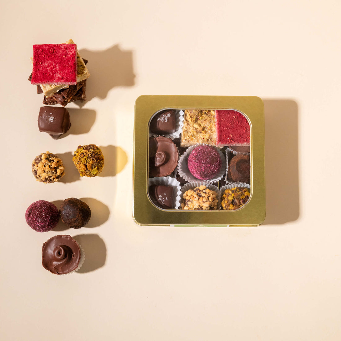 An assortment of gourmet chocolate truffles and molded chocolates arranged beside an open square gold tin showing the variety inside, on a warm beige background.
