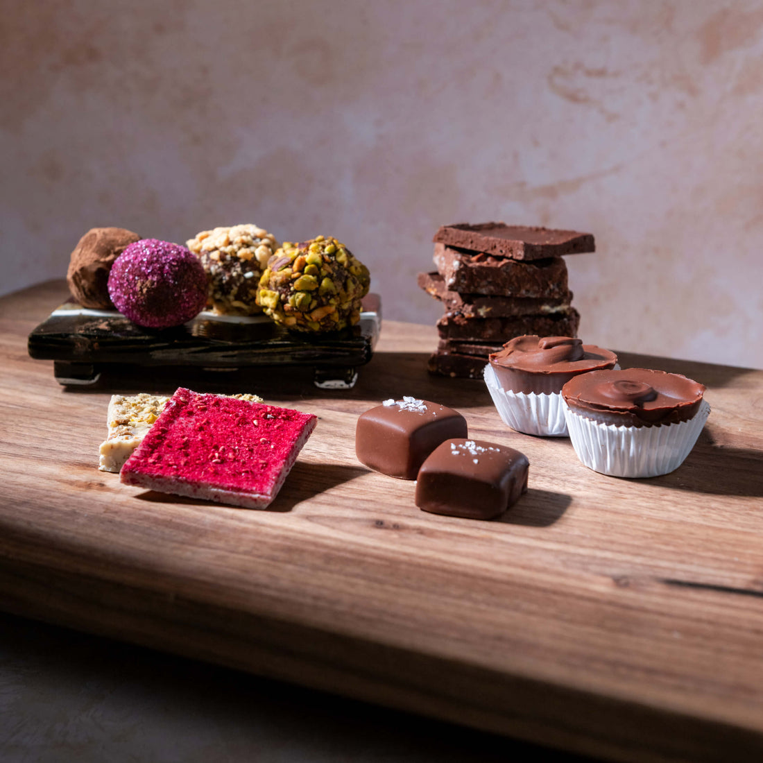 An assortment of gourmet chocolates and truffles displayed on a wooden board against a textured beige background. The collection includes milk and dark chocolate squares, chocolate-covered cupcakes, and colorful truffles, including one with a pink raspberry topping and one with a pistachio coating.