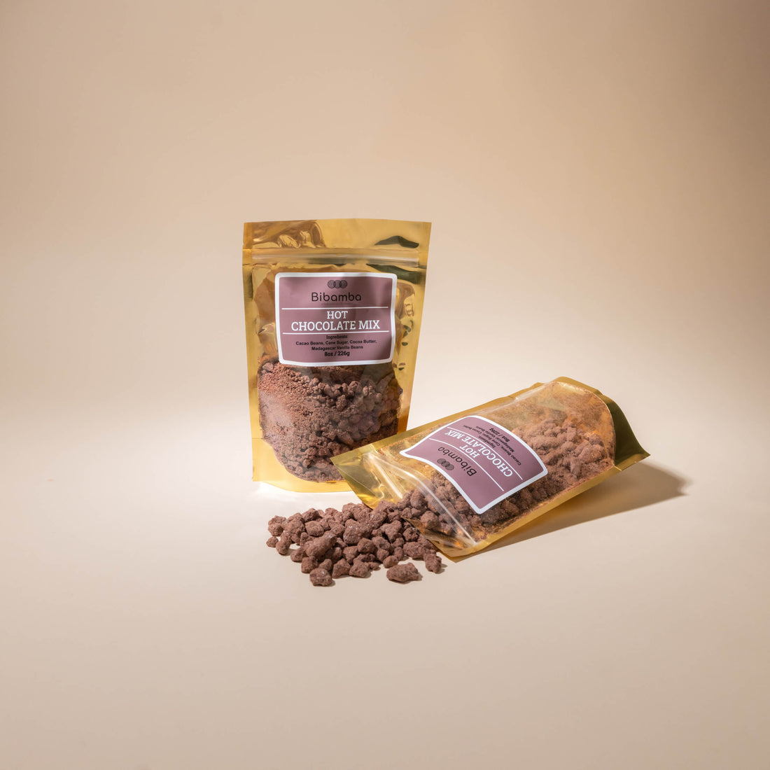 Two stand-up pouches of Bibamba hot chocolate mix on a warm beige studio background, with one pouch upright and one lying on its side. A small pile of dark brown chocolate powder sits in front.