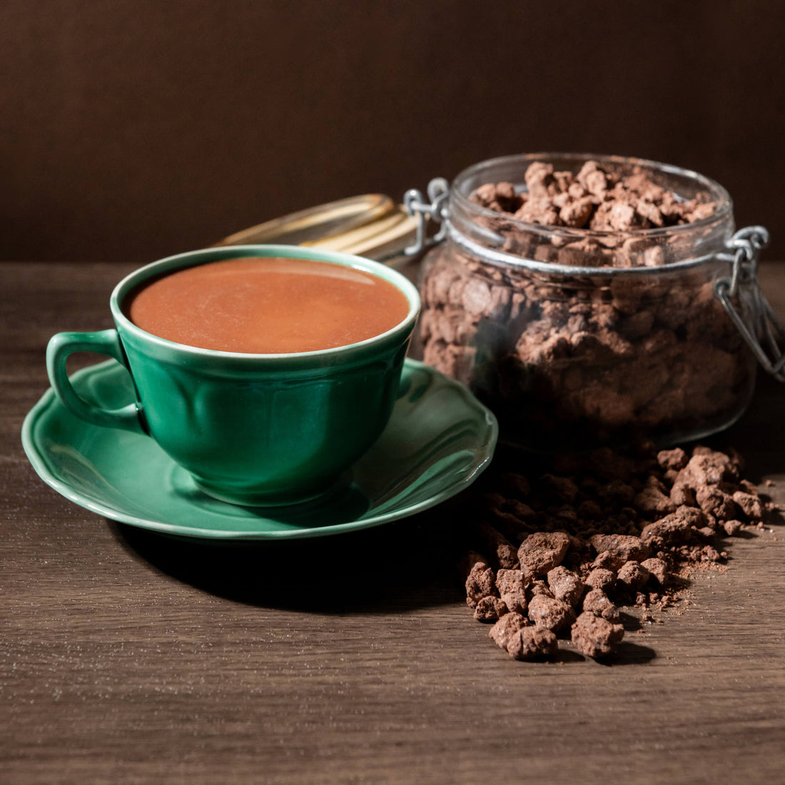 A green ceramic cup filled with Bibamba hot chocolate sits on a matching saucer beside a glass jar of Bibamba hot chocolate mix.