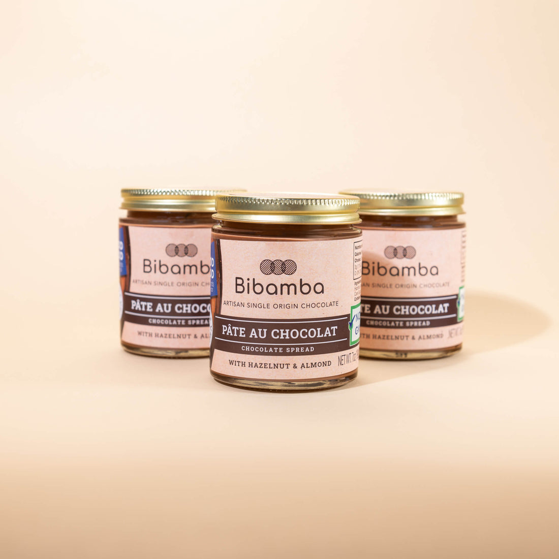 Three glass jars of Bibamba chocolate spread arranged in a row on a warm beige background, with the center jar in sharp focus and the rear jars softly blurred.