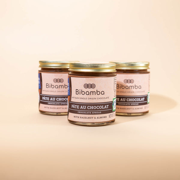 Three glass jars of Bibamba chocolate spread arranged in a row on a warm beige background, with the center jar in sharp focus and the rear jars softly blurred.