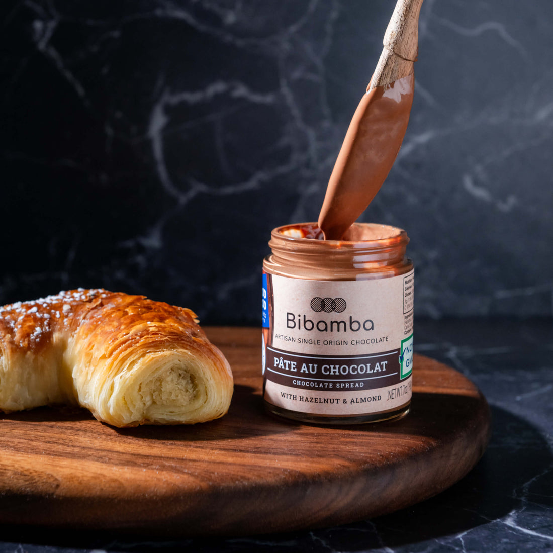 A jar of Bibamba chocolate spread sits on a wooden board next to a pastry, with a wooden spoon lifting chocolate spread from the open jar against a dark marble background.