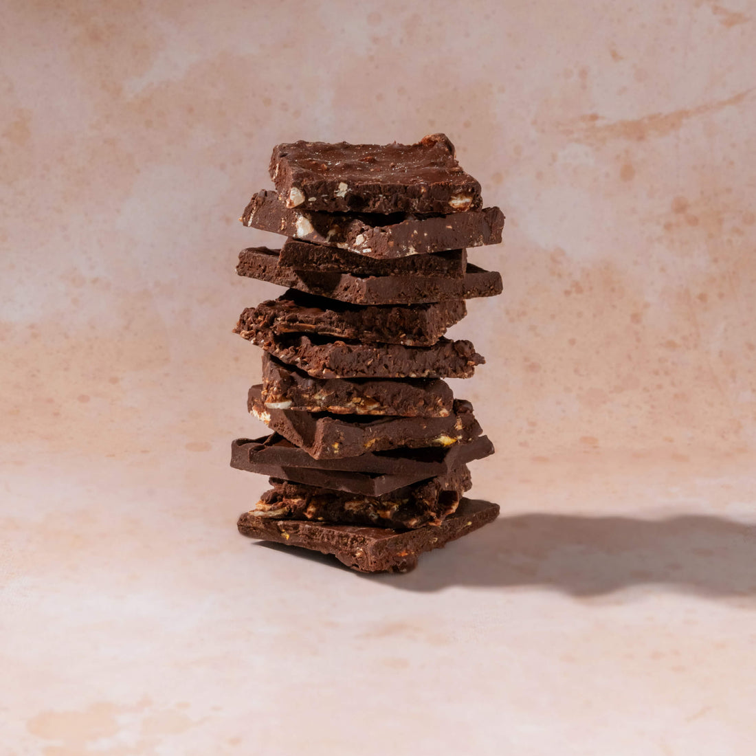 A stack of rustic dark chocolate bark pieces with visible nuts and white chocolate pieces, standing upright on a light beige textured surface.