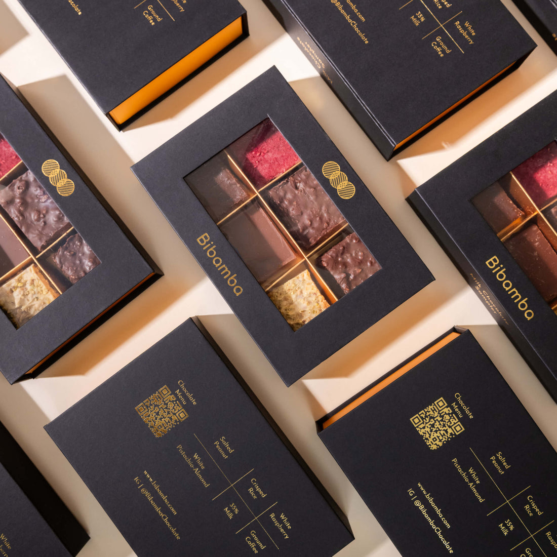 Several black gift boxes of Bibamba milk chocolate samplers arranged in a flat-lay pattern on a light beige background. The boxes feature a clear window displaying an assortment of chocolate bars, with gold foil branding and details.