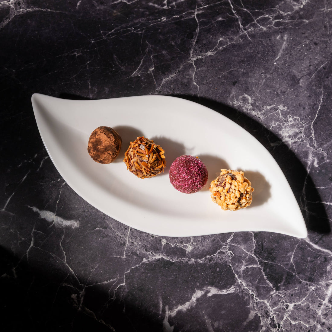 Four gourmet Bibamba chocolate truffles in arranged on a white leaf-shaped ceramic plate, set against a dark gray marble background.