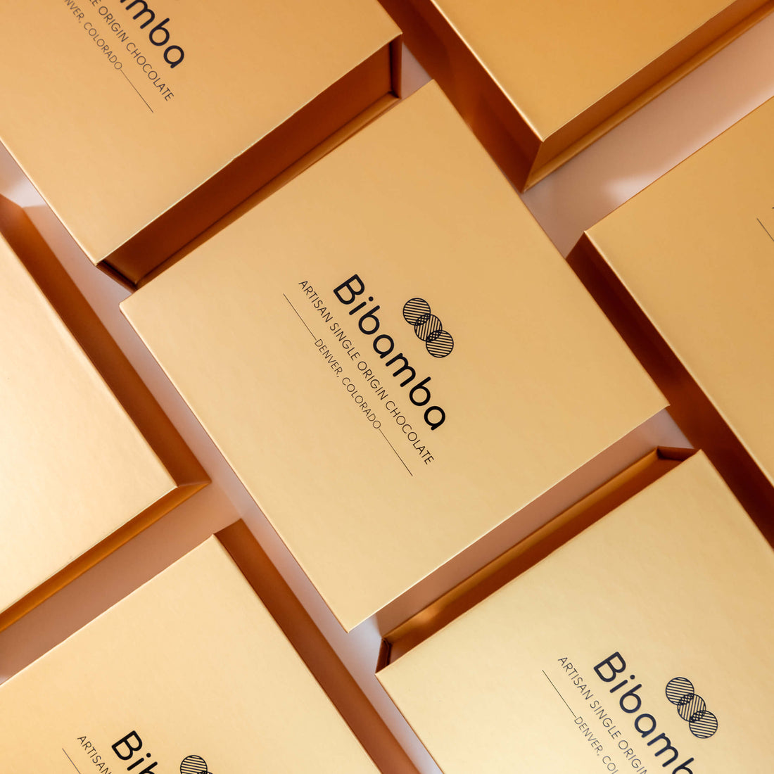 Multiple gold-colored rectangular boxes arranged in a diagonal grid, each printed with the brand name 'Bibomba' and product text for artisan single-origin chocolate.