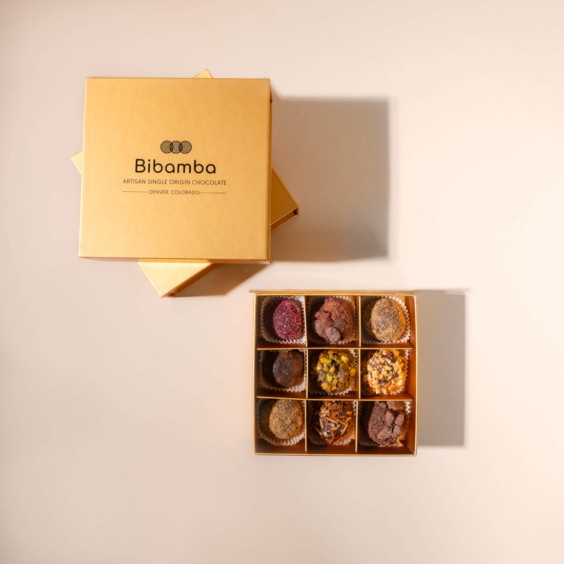 An open gold box of Bibamba artisan single-origin chocolate truffles displayed next to its gold lid on a warm beige background.